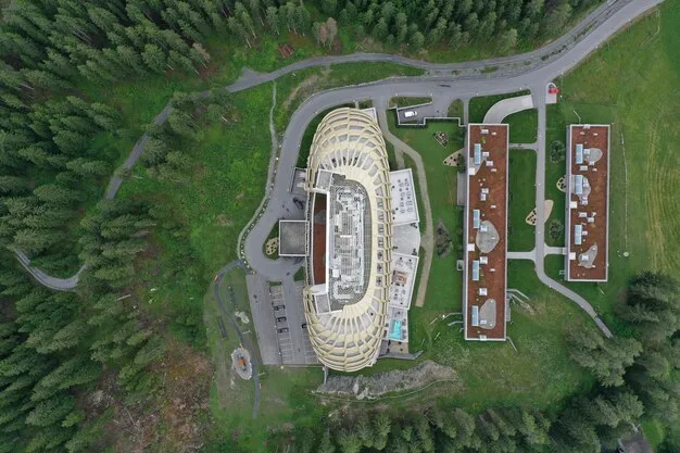 An aerial view of a newly constructed MLS stadium surrounded by parks, residential buildings, and commercial spaces. The stadium's design incorporates sustainable elements, such as solar panels and green roofs, blending into the urban environment.